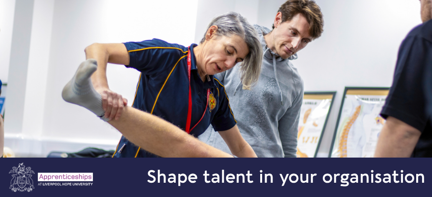 A female Physiotherapy lecturer is gripping the leg of a patient and demonstrating a technique to a male student who is stood directly behind her.